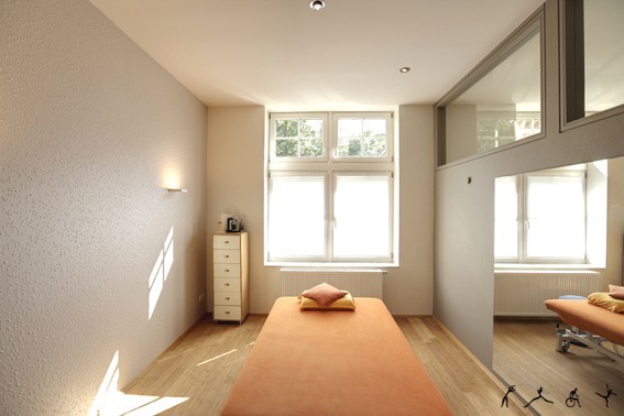 Heller Behandlungsraum mit Massageliege, Fenster und moderner, barrierefreier Einrichtung.