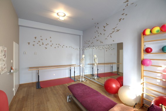 Heller Physiotherapieraum mit Sprossenwand, Gymnastikbällen, Spiegel und Skelettmodellen.
